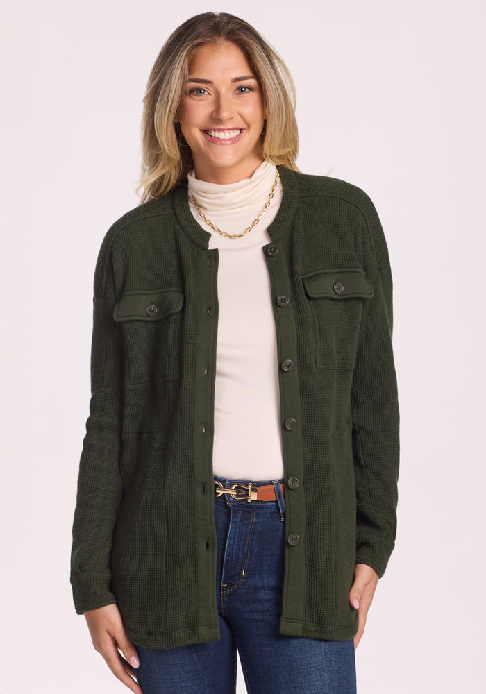 A smiling woman with long blonde hair wears the Woolx Oaklynn Waffle Shacket in Driftwood Fern over a white turtleneck and blue jeans, standing against a plain white background. 