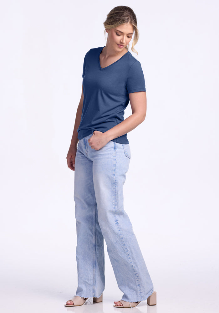 A woman stands against a white background, wearing the Woolx Junie V Neck in Starry Night, light blue wide-leg jeans, and beige heeled sandals. She looks down with one hand in her pocket.