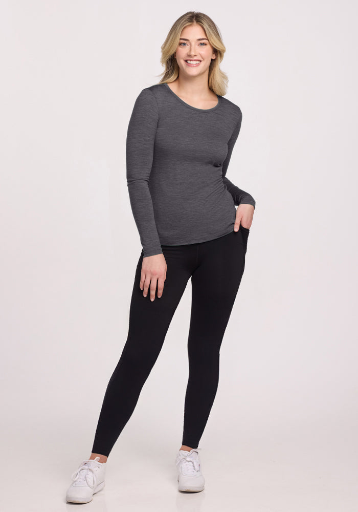 A woman smiles, wearing the Woolx Remi Long Sleeve T-Shirt in Cinder Melange—an odor-resistant, moisture-wicking Merino wool top—paired with black leggings and white sneakers, one hand in her pocket against a plain light background.