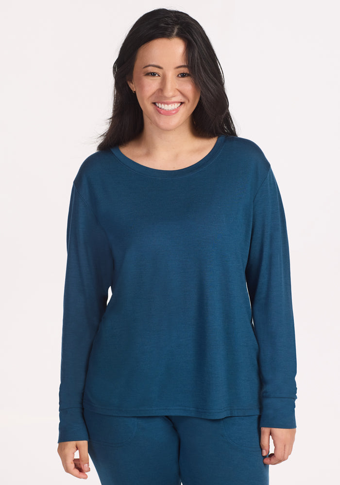A woman with long dark hair smiles at the camera, wearing the Woolx Ashlie Lounge Top in Majolica Blue, against a plain light background. 