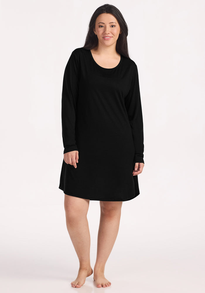A woman with long dark hair stands barefoot and smiles against a plain light background, wearing the Woolx Lucille Long Sleeve Nightgown in black, a simple merino wool style that falls above the knees. 