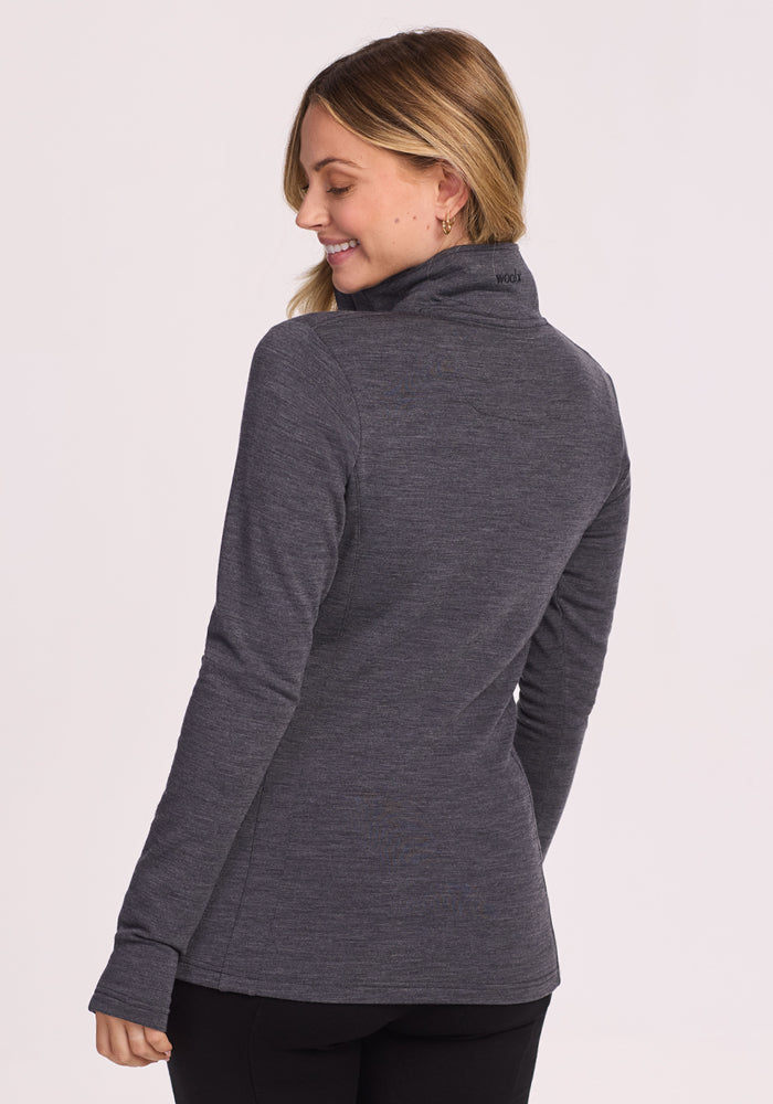 A woman with blonde hair wears the Woolx Finley Zip Up Sweatshirt in Cinder Melange, standing against a light background and smiling over her shoulder.