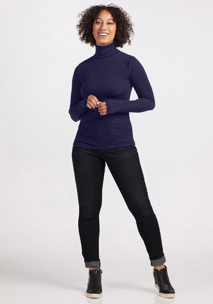 A person with curly hair smiles against a plain light background, wearing the Woolx Sage Ribbed Turtleneck in Midnight Sapphire, paired with dark jeans and black high-top shoes.