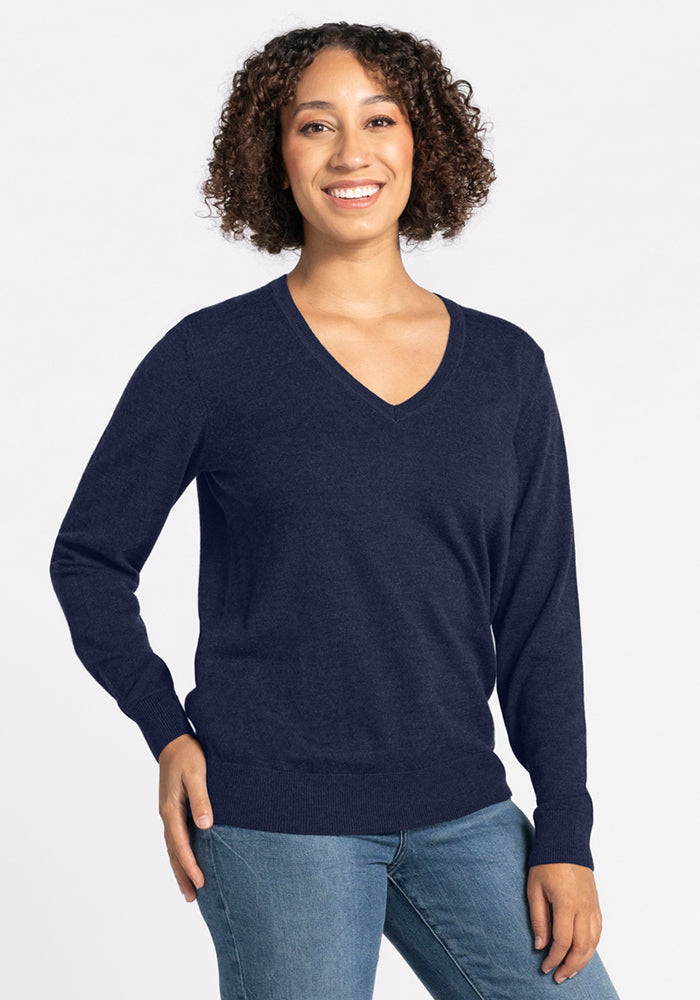 A person with curly hair, wearing the Woolx Francesca Sweater in Storm Cloud and blue jeans, stands smiling against a plain light background, with one hand by their side and the other on their hip. 
