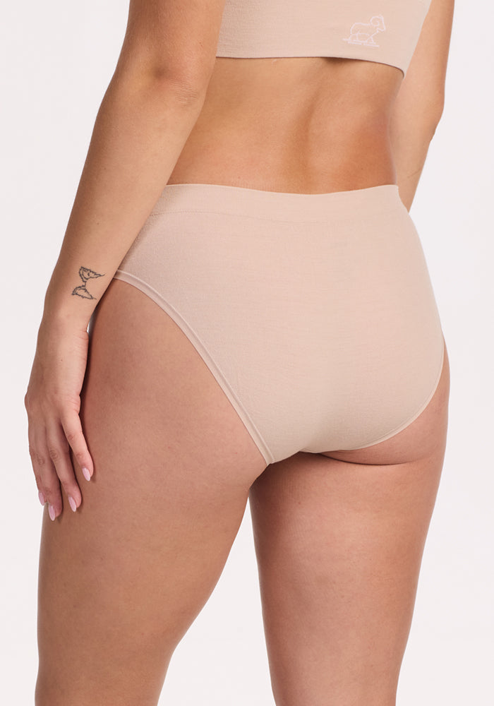 A person wears the Woolx Maeve Bikini in Sand, paired with a matching bra. They stand slightly turned, revealing a small bird tattoo on their left forearm against a plain white background.