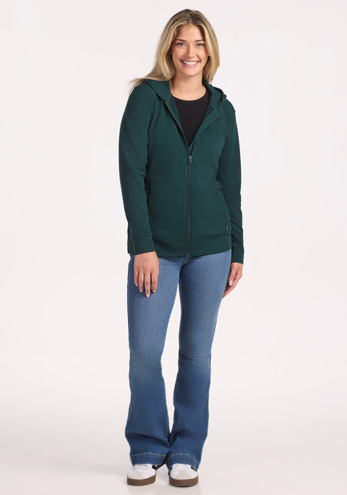 A woman with long blonde hair smiles while standing against a white background. She is wearing the Woolx Darby Zip Up Hoodie in Deep Timber over a black shirt, with blue jeans and white sneakers.