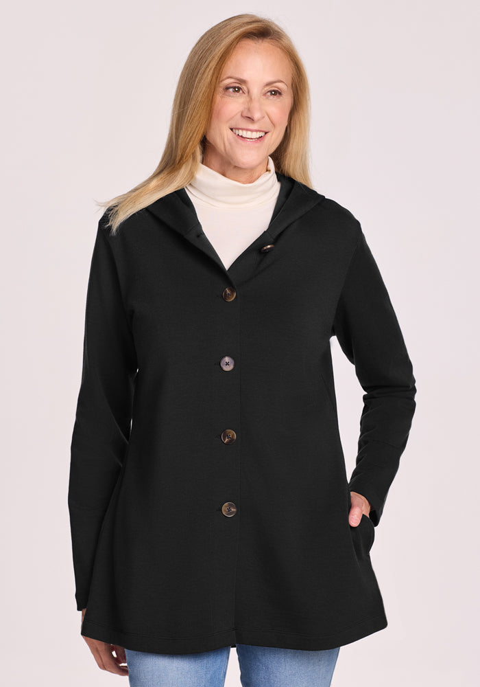 A smiling woman with long blonde hair wears the Woolx Edna Hooded Cardigan in black over a white turtleneck and blue jeans, standing with one hand in her pocket against a light background. 