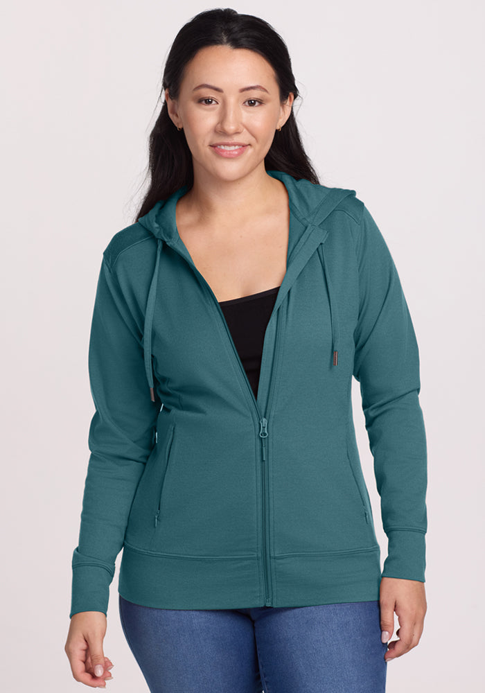 A woman with long dark hair smiles, facing forward in the Woolx Ryann Hoodie - Tidepool over a black top and blue jeans against a light background—a must-have travel essential. 