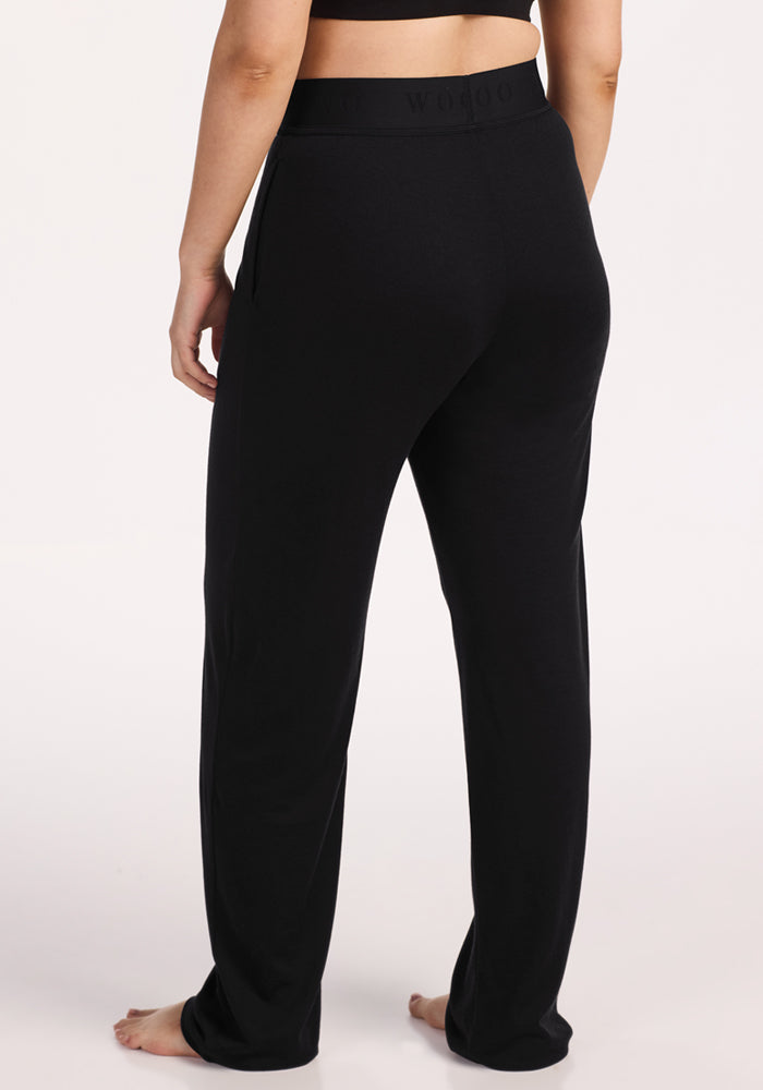 A person stands barefoot on a white background, shown from the back, wearing Woolx Maya Lounge Pants in black and a matching black crop top.