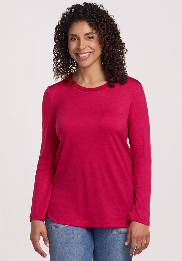 A woman with curly hair smiles in the Woolx Hazel Tunic - Poppy Spark, a bright pink, lightweight merino wool top paired with blue jeans. She stands against a plain light background, showcasing the tunic's stylish, temperature-regulating fabric. 