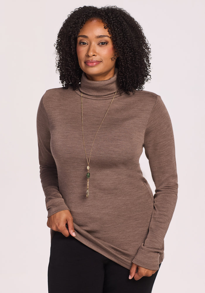 A woman with curly hair smiles gently, wearing the Woolx Peyton Turtleneck in Simply Taupe, a long green-accented necklace, and black pants. She stands against a plain light background, holding the hem of her sweater. 