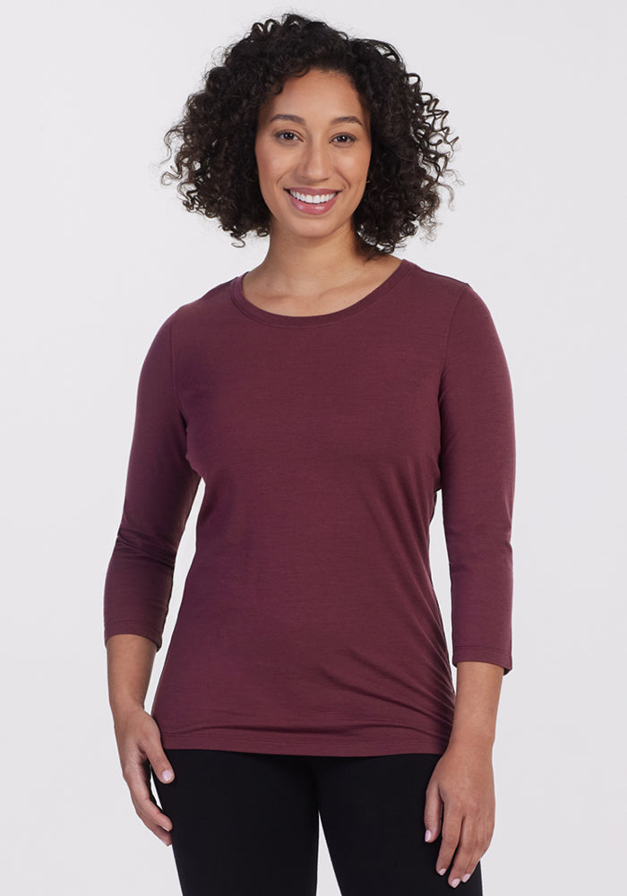 A woman with curly hair smiles, wearing the Woolx Jenny 3/4 Sleeve Crew Neck in Grape Harvest and black pants, standing against a plain light background. 