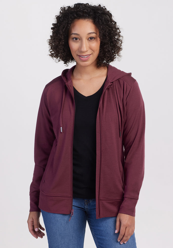 A woman with curly hair smiles at the camera, wearing the Woolx Ryann Hoodie in Grape Harvest over a black shirt and blue jeans, standing against a plain white background. 