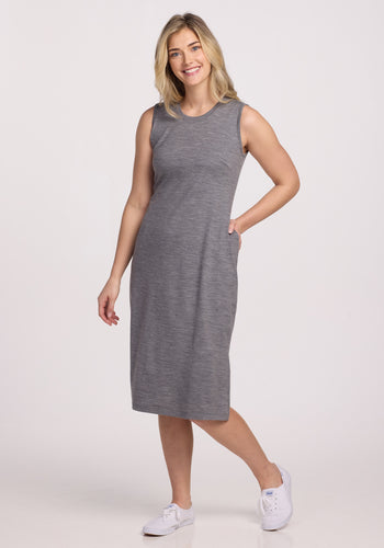 A blonde woman smiles while wearing a Woolx Cassie Dress in Graphite Heather and white sneakers, standing with one hand in her pocket against a plain white background. | Ariana is 5’11”, wearing a size S