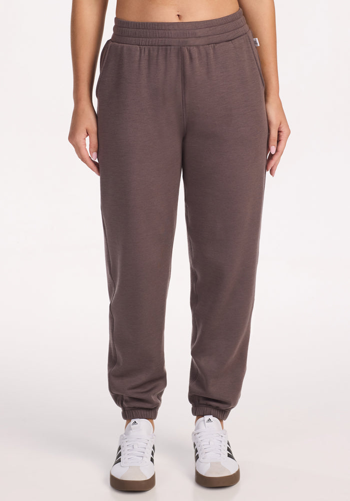 A person in high-waisted, plum truffle Woolx Parker Sweatpants with an elastic waistband and cuffs, paired with white sneakers, stands against a white background, enjoying temperature regulation and moisture-wicking comfort. 
