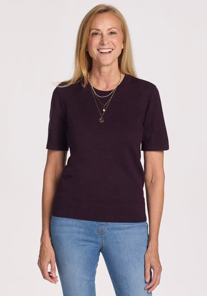 A smiling woman with long blonde hair wears the Woolx Anna Crew Sweater in Deep Plum, layered gold necklaces, and blue jeans, standing against a plain white background. 