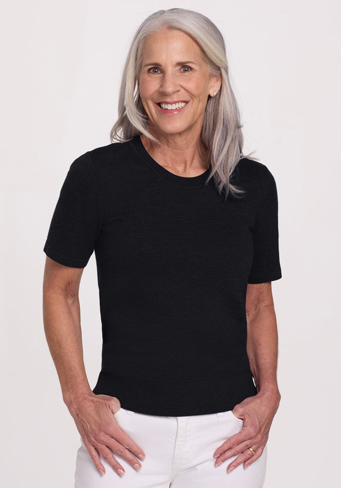 Smiling older woman with long gray hair wears the Woolx Anna Crew Sweater in black and white pants, standing against a plain light background. 