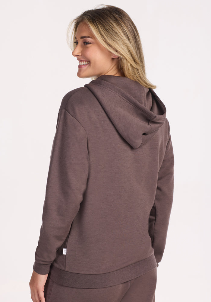 A person with light brown hair smiles over their shoulder, wearing the Woolx Avery Hoodie in Plum Truffle. The shot, taken from behind, features a plain, light background.