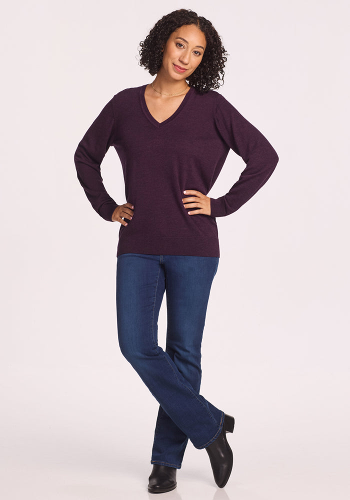 A woman with curly hair stands confidently with hands on hips, smiling in the Woolx Francesca Sweater in Deep Plum, blue jeans, and black ankle boots against a plain white background.