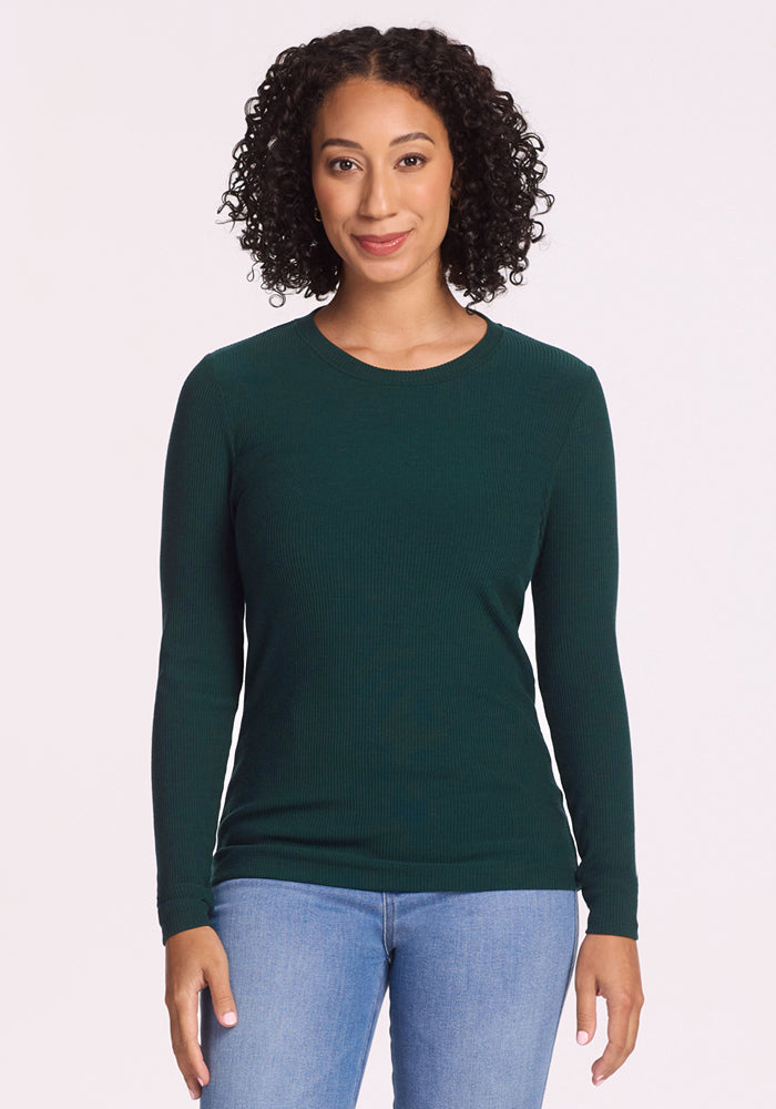 A woman with curly hair wears the Woolx Hadley Ribbed Crew in Deep Timber and light blue jeans, standing facing forward and smiling slightly against a plain light background. 