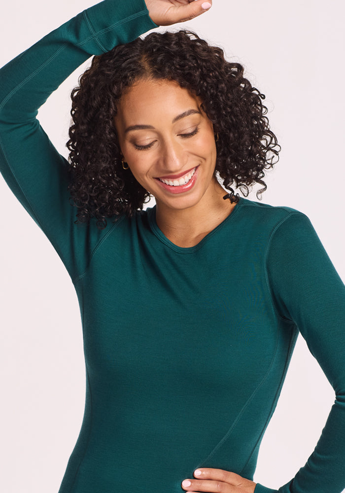 A woman with curly hair smiles with her eyes closed and one arm raised, wearing the Woolx Hannah Long Sleeve Top in Riverbank Green against a light background.