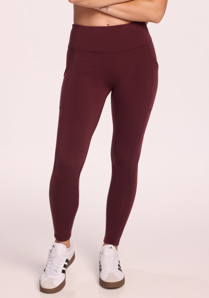 A person models Woolx Piper Pocket Leggings in Toasted Garnet, paired with white sneakers. The high-waisted leggings feature side pockets. The image is cropped from midriff to feet against a light, neutral background. 