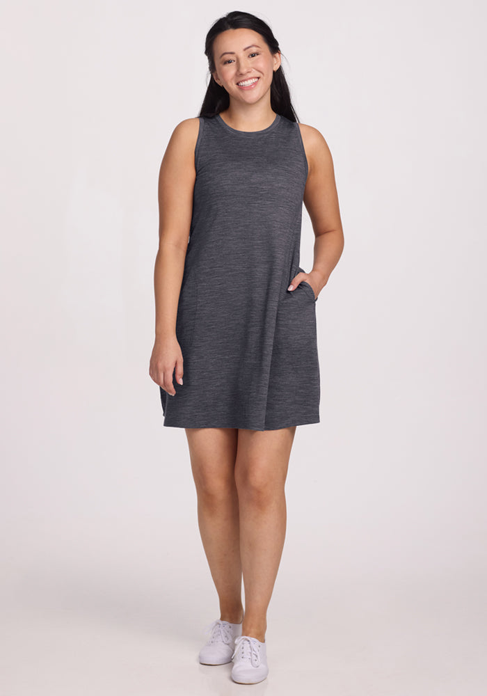 A woman with long dark hair smiles with one hand in her pocket, wearing the Woolx Clara Dress in Cinder Melange and white sneakers, posed against a plain light background. 