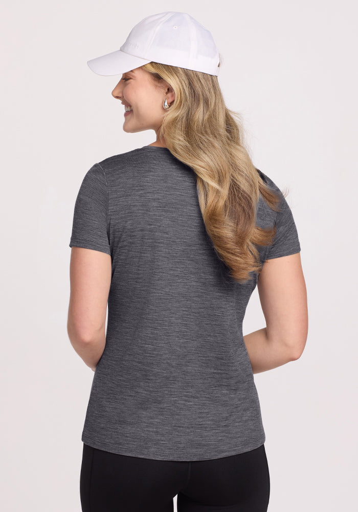 A woman with long, wavy blonde hair wears a white baseball cap, black pants, and the Woolx Junie V Neck tee in Cinder Melange. She stands with her back to the camera, smiling against a plain light background.