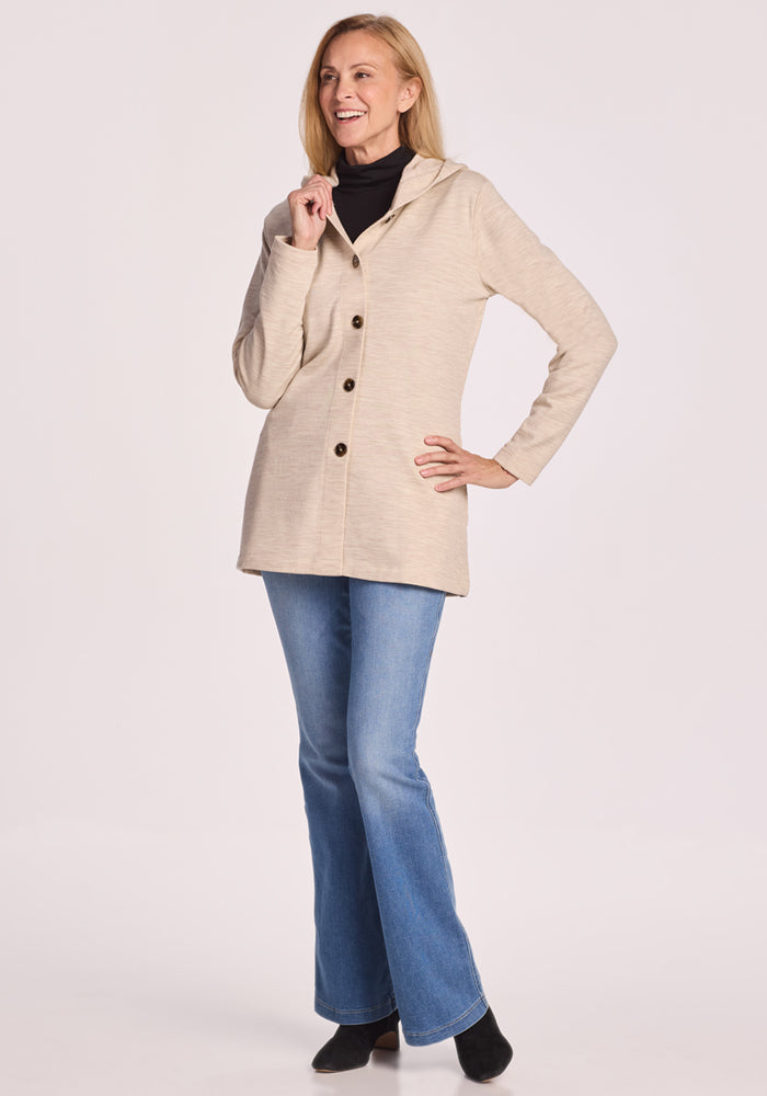 A woman smiles in the Woolx Edna Hooded Cardigan in Cream Heather, paired with blue jeans and black shoes, set against a plain light background.