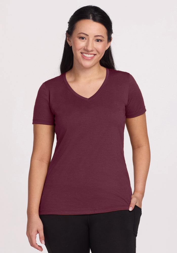 A woman with long dark hair, wearing the Woolx Junie V Neck in Grape Harvest and black pants, smiles with one hand in her pocket against a plain light background. 