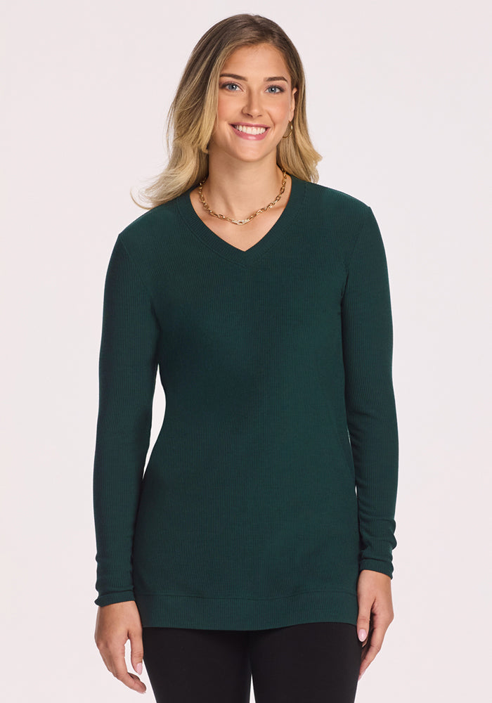 A woman with light brown hair smiles at the camera, wearing black pants and the Woolx Evie Ribbed Tunic in Deep Timber—a cozy women's long-sleeve Merino wool V-neck—while standing against a plain white background. 