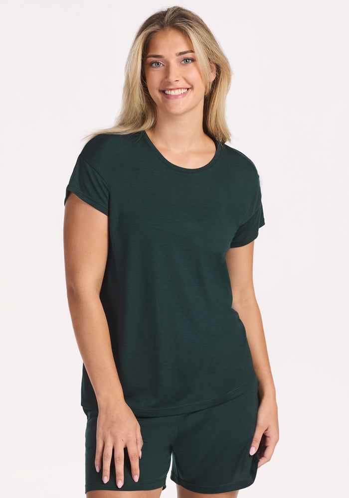 A woman with long blonde hair smiles while wearing the Woolx Billie Lounge Tee in Deep Timber, paired with matching shorts. She stands against a plain light background.