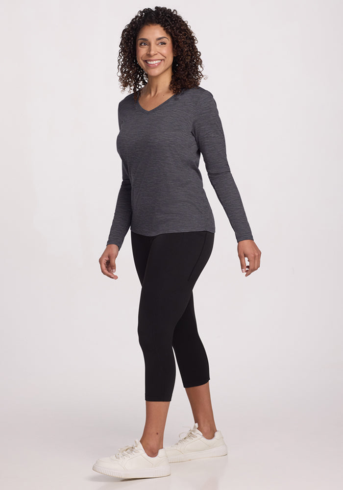 A woman with curly hair, smiling as she walks against a plain light background, wears the Woolx Millie Long Sleeve in Cinder Melange, paired with black leggings and white sneakers.