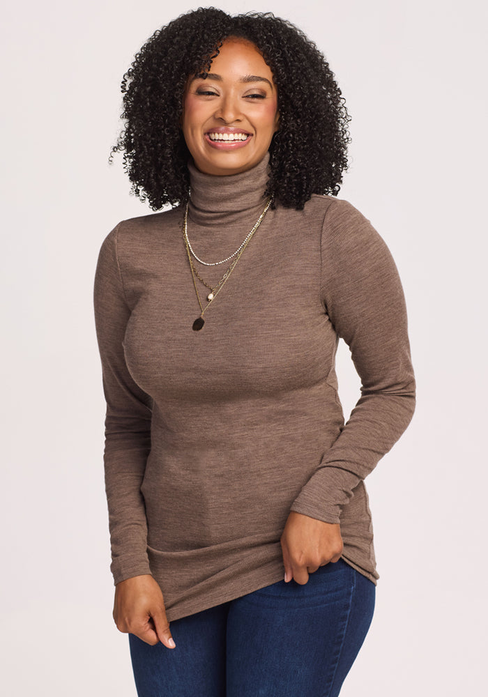 A smiling woman with curly hair wears the Woolx Sage Ribbed Turtleneck in Simply Taupe, layered gold necklaces, and blue jeans, standing against a plain light background. 
