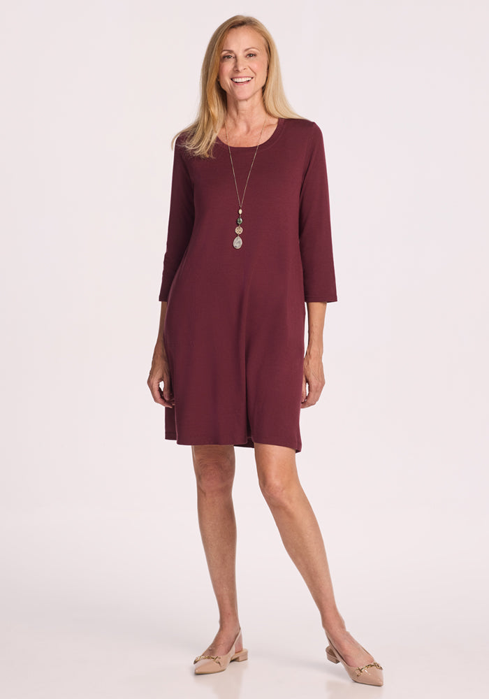 A woman with long blonde hair models the Woolx Rissa A Line Dress in Toasted Garnet, a maroon Merino wool knee-length dress with 3/4 sleeves—versatile for fall/winter layering or chic travel, styled with a necklace and beige flats. 