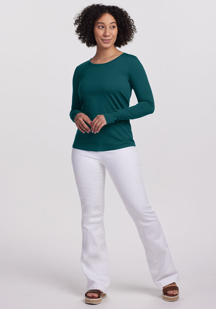 A woman with curly hair stands against a plain background, wearing the Woolx Remi Long Sleeve T-Shirt in Riverbank Green, white pants, and tan platform sandals. She faces slightly to the side with her hands together in front of her.