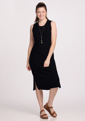 A person with long hair is smiling while wearing the Iris Ribbed Dress in black by Woolx, accessorized with a long necklace. Their hair is tied back as they stand against a plain background, pairing the look with brown sandals. | Lilly is 5’9”, wearing a size S