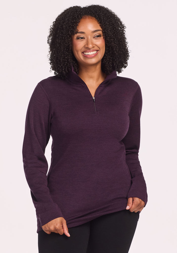 A woman with curly hair smiles while wearing the Woolx Brooke 1/4 Zip in Deep Plum and black pants, standing against a plain light background. 