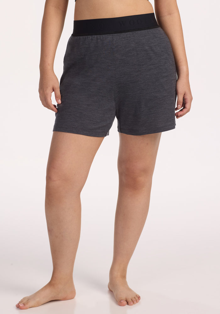 A person stands barefoot on a light background, wearing Woolx Poppy Lounge Shorts in Cinder Melange, featuring a black elastic waistband. Their arms are relaxed by their sides and their head is out of frame. 