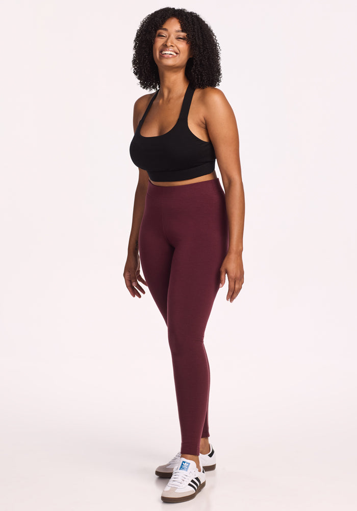 A woman with curly hair smiles while standing, wearing a black sports bra, Woolx Stella Leggings in Toasted Garnet, and white Adidas sneakers against a plain white background.