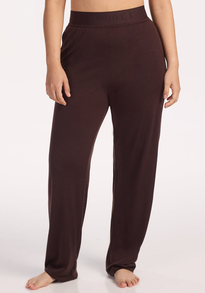A person stands barefoot in loose-fitting Maya Lounge Pants in Chocolate Plum by Woolx, featuring an elastic waistband with "WOOLX" branding, against a plain white background. 