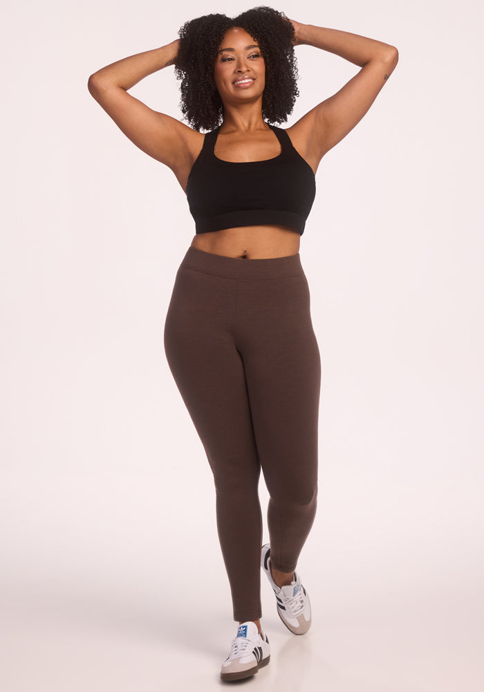 A woman with curly hair wears Woolx Stella Tall - Bracken DuraLite leggings, a black sports bra, and white sneakers, smiling with her hands behind her head against a plain white background.