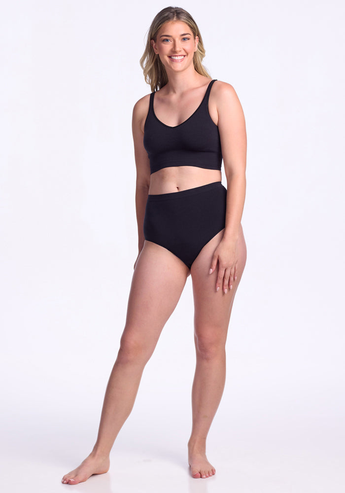A woman with long blonde hair stands barefoot and smiling in a black sports bra and Woolx Mabel Full Coverage Underwear - Black, offering seamless comfort, against a plain white background.