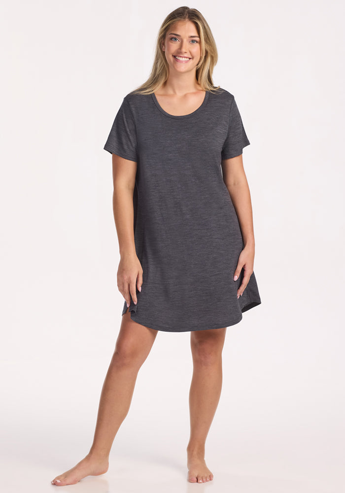 A smiling woman stands barefoot in the Woolx Desi Nightgown in Cinder Melange—a short-sleeved, dark gray Merino wool sleepwear with a relaxed fit—set against a plain light background. 