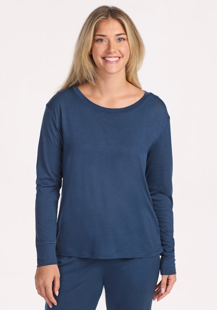 A woman with long blonde hair smiles at the camera, wearing the Woolx Ashlie Lounge Top in Starry Night—moisture-wicking Merino wool. She stands against a plain light background. 
