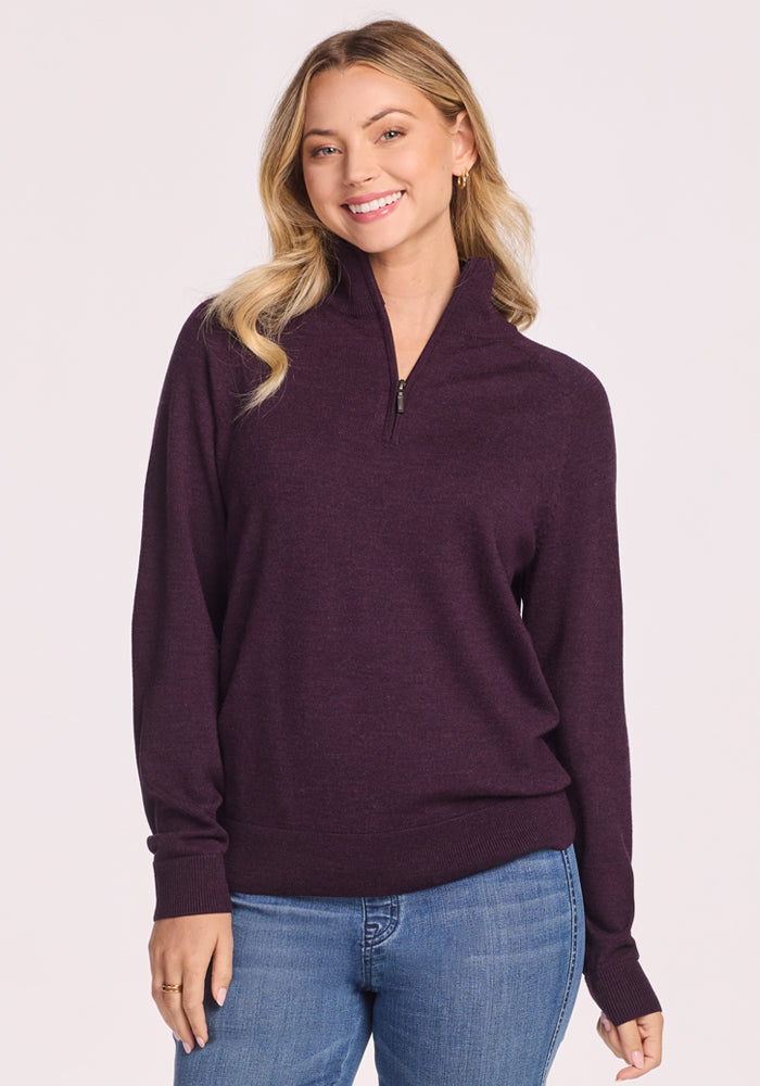 A woman with long blonde hair, dressed in the Woolx Collins Half Zip Sweater in Deep Plum and blue jeans, smiles while standing against a plain light background. 