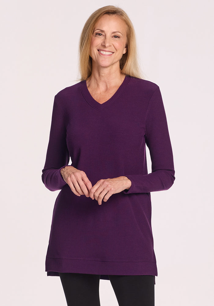 A woman with long blonde hair smiles in the Woolx Evie Ribbed Tunic in Majestic Purple and black leggings, standing against a plain light background. Its temperature-regulating Merino wool keeps her comfortable all day. 