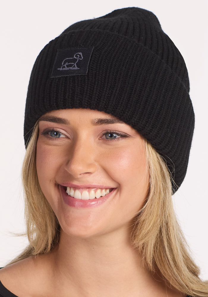 A smiling person wears the Woolx Ria Chunky Beanie in black, featuring a small patch with a simple duck line drawing on the front. This cozy winter accessory stands out against the plain white background.