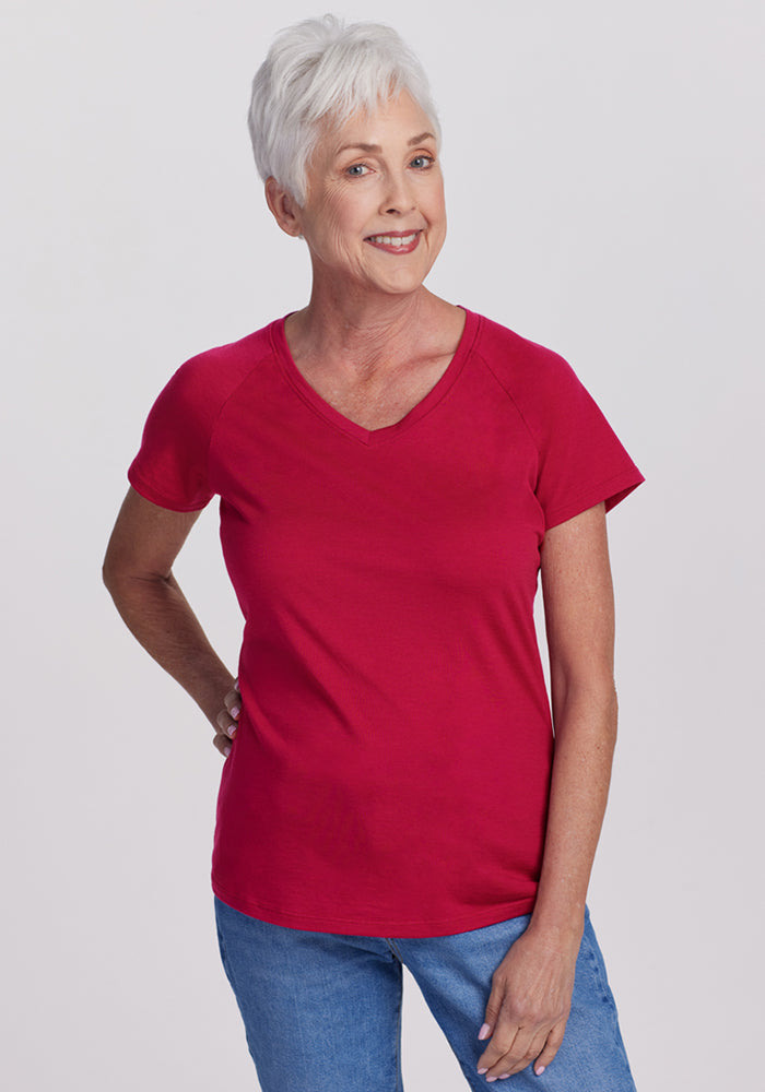 A smiling older woman with short gray hair wears the Woolx Mia V Neck in Poppy Spark and blue jeans, standing with one hand on her hip against a plain light background. 