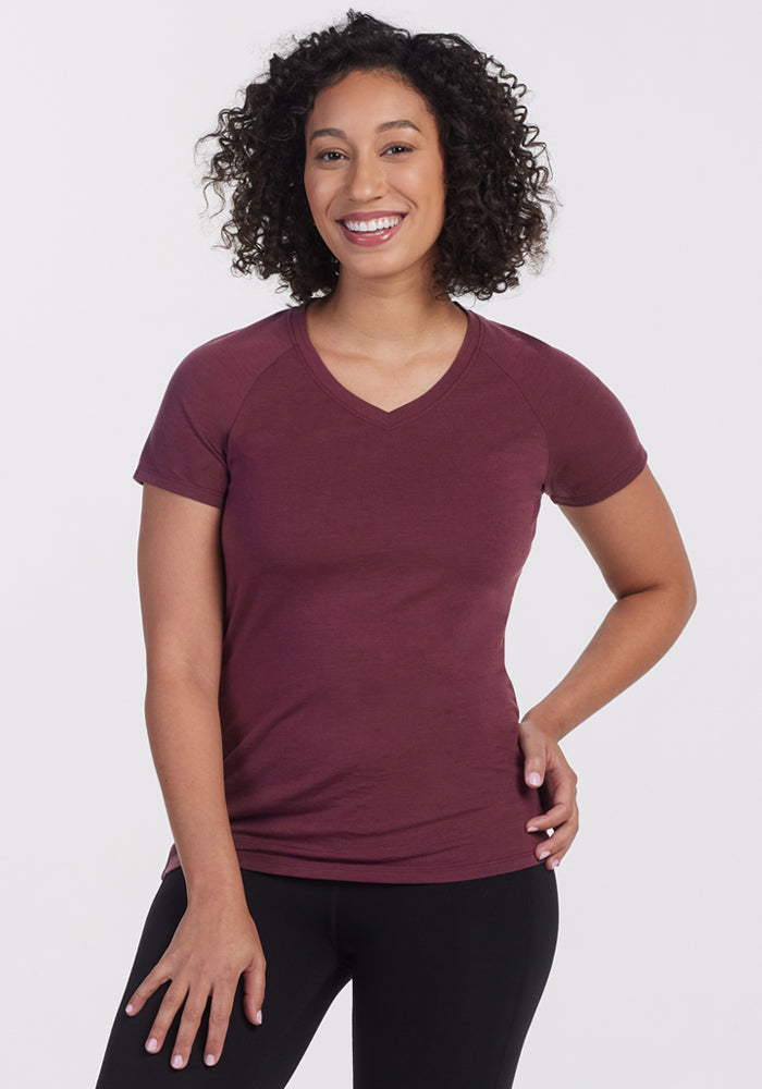 A woman with curly hair smiles at the camera, wearing the Woolx Mia V Neck in Grape Harvest and black pants. She stands against a plain white background with one hand on her hip. 